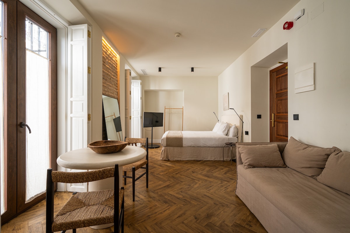 A modern studio apartment features a spacious seating area with a neutral-colored sofa, a round dining table, and a full-length mirror. A large bed is positioned against a wall with warm wooden tones, while natural light enters through double doors that lead to a small balcony.