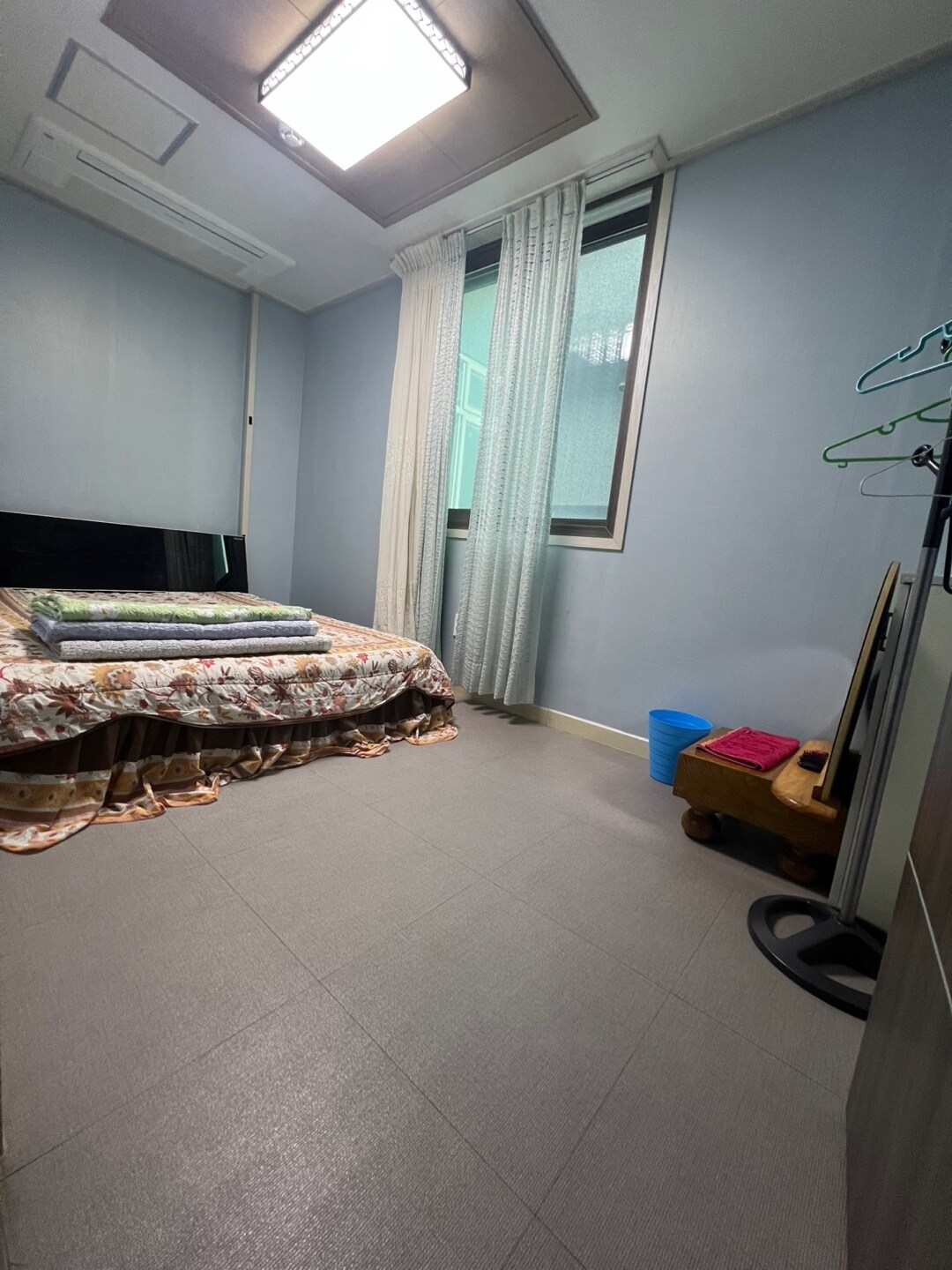 A bright room features a spacious bed with a floral bedspread, complemented by a folded blanket. Natural light enters through a large window, which is dressed with sheer curtains. A chair and a small blue bin are also visible, along with a clothing rack.