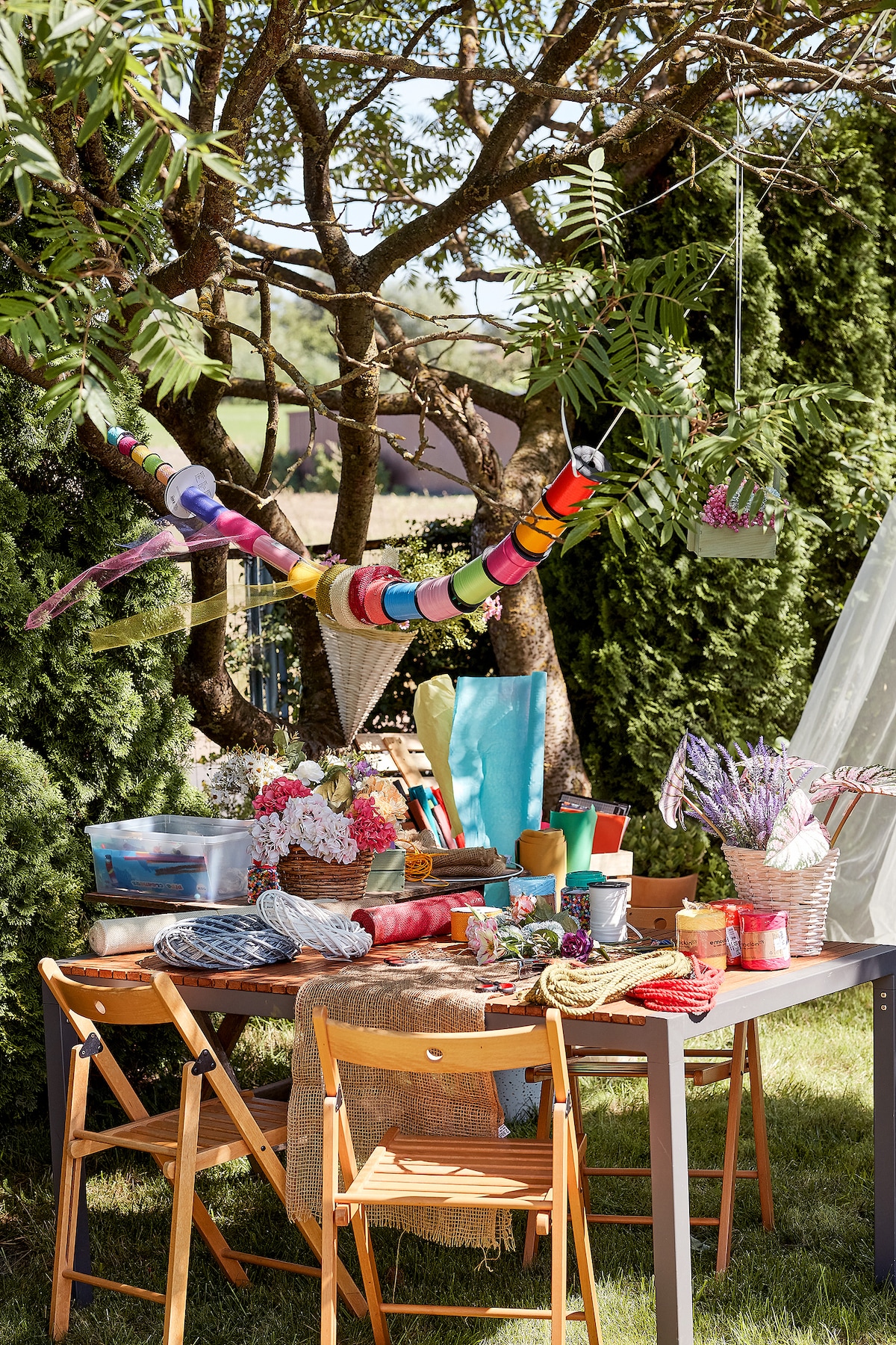 A charming outdoor workspace is set up under a tree, featuring a table surrounded by wooden chairs. Colorful craft supplies, including ribbons and flowers, are organized in baskets and containers, creating an inviting creative area. Lush greenery adds to the serene environment.