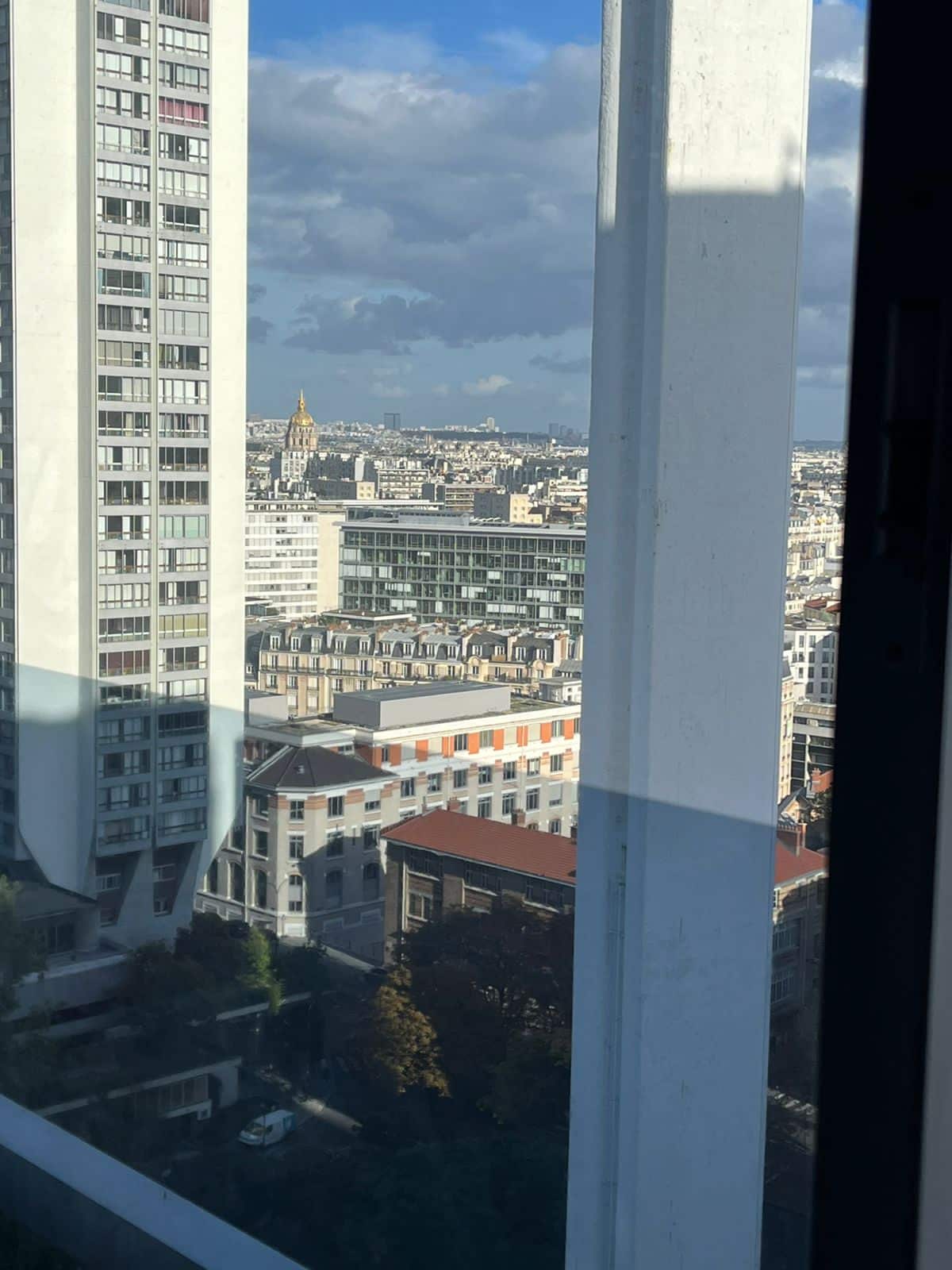 A panoramic view of Paris is framed by the window, revealing a mix of modern and classic buildings below. The iconic Paris skyline, including a glimpse of a prominent golden structure, is visible under a partly cloudy sky.