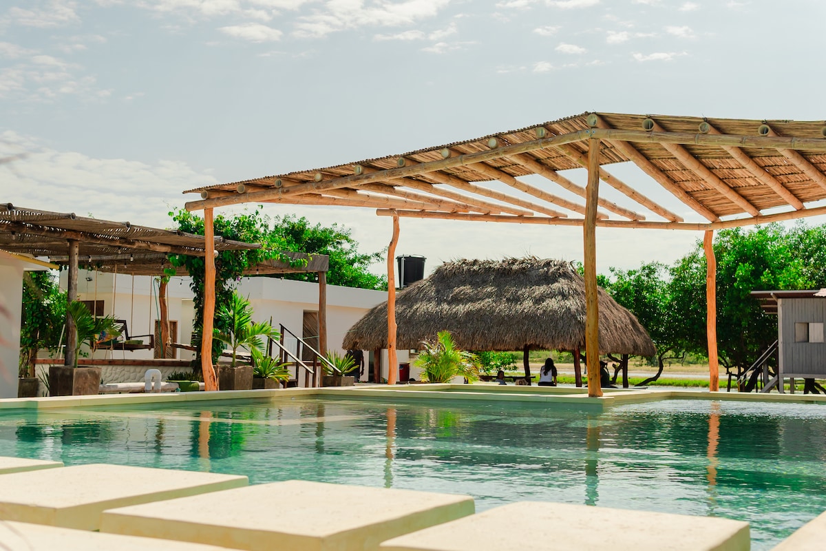 An inviting outdoor pool is surrounded by a spacious wooden deck with a shaded area featuring a thatched roof. Lush greenery is visible in the background, enhancing the serene environment. Clear skies and soft sunlight create a peaceful atmosphere around the swimming area.