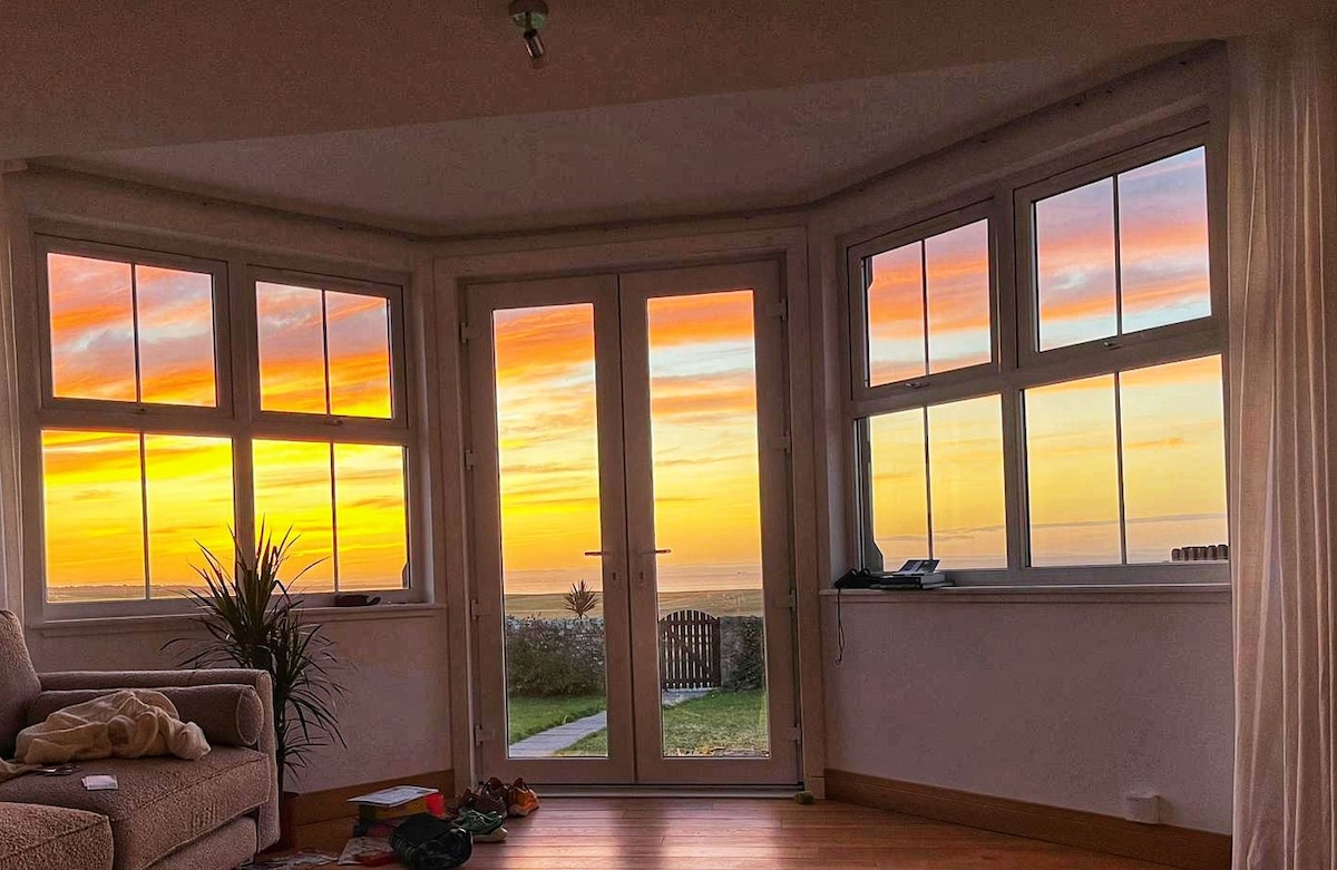 Large windows frame a vibrant sunset, revealing hues of orange and pink across the sky. Natural light floods the room, complemented by a potted plant and soft furnishings. A wooden floor leads to a grassy area outside, enhancing the connection to nature.