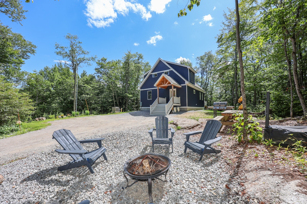 A fire pit area is surrounded by four adirondack chairs, positioned on a gravel surface. The modern lake house is visible in the background, framed by trees under a clear blue sky. The inviting outdoor space is ideal for gathering and relaxation.