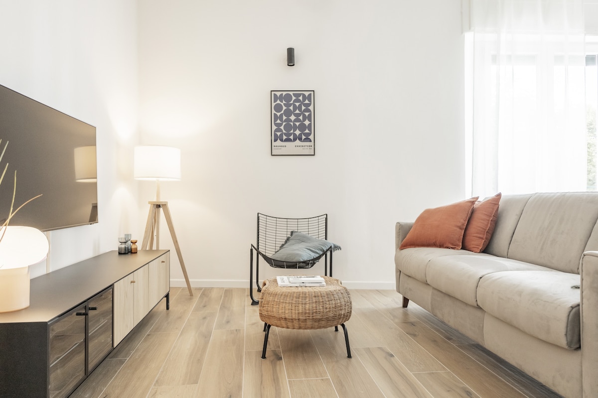 A light-filled living area features a soft-colored sofa adorned with orange accents. A sleek entertainment unit is positioned against a neutral wall, complemented by a standing lamp and a contemporary artwork. A cozy seating option is arranged nearby on a wooden floor, enhancing the welcoming feel.