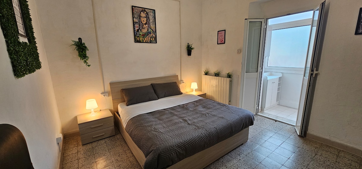 The spacious bedroom features a large bed with neutral bedding, complemented by bedside lamps on either side. A window offers natural light, and a doorway leads to an adjoining area. Green plants are placed on the walls, adding a touch of nature to the setting.