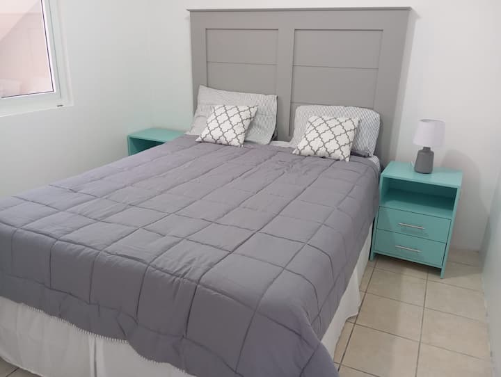 Cama Quee ubicada en el primer nivel  en un habitación con sus almohadass, almohadones, mesas y lámpara de noche. Cuenta con una cabecera minimalista, ventana con cortina y puerta con seguro interno.