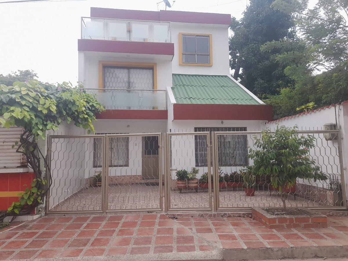 The image displays a three-level house surrounded by greenery. A gated entrance leads to a paved area with several potted plants. The upper floor features a balcony with a green roof and large windows, while the ground level has a welcoming façade with light colors.