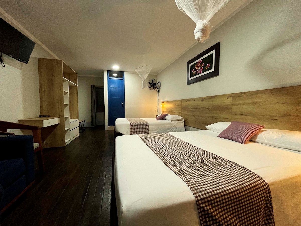 A spacious room features two beds covered with white linens, accented by checkered throws. A wooden headboard serves as a backdrop, while a wall-mounted television and a decorative piece add character. A study area with a desk and chair, along with a wardrobe, is visible.