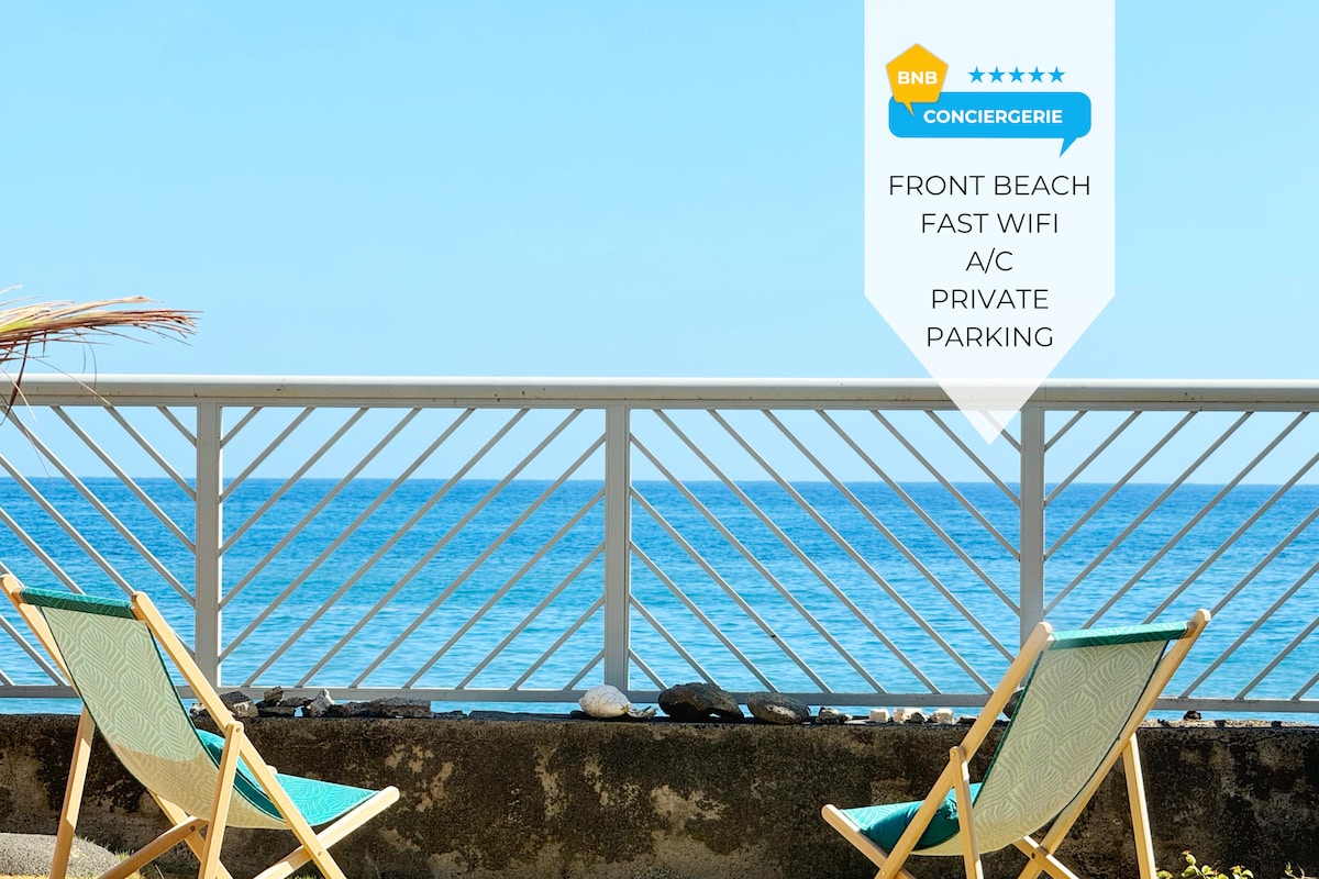 Two wooden lounge chairs are positioned on a stone patio, facing a serene beach view. A railing separates the seating area from the calm blue ocean, enhancing the peaceful ambiance of the location.