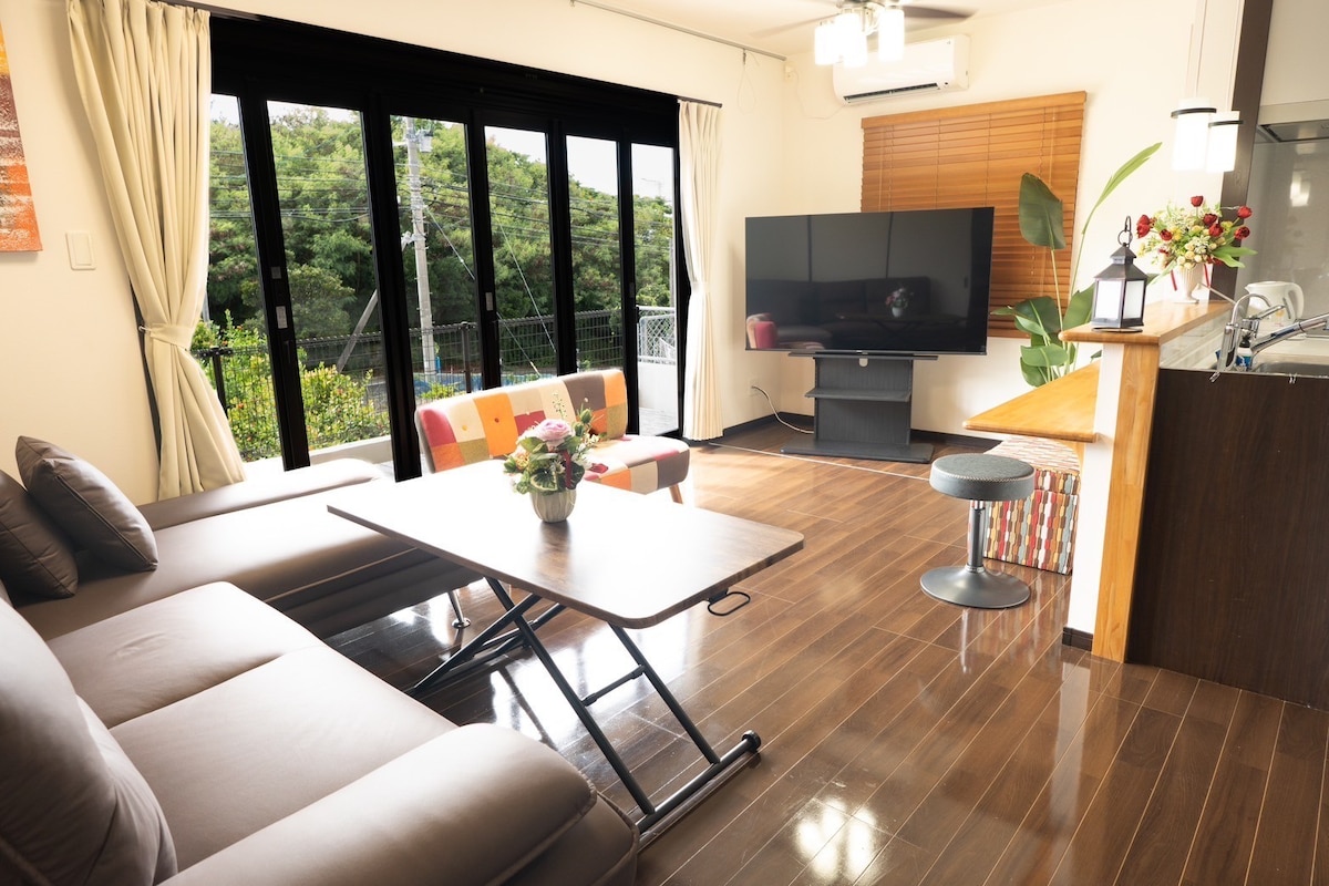 The living area showcases a comfortable sofa and a coffee table, surrounded by large glass doors that invite natural light. A 65-inch television stands across from the seating, while a dining area with a kitchen counter is visible in the background.