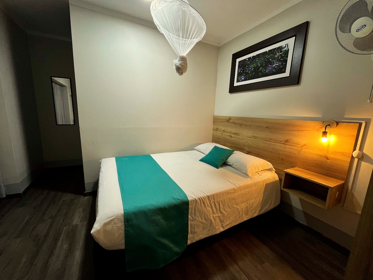 A comfortable sleeping area features a spacious bed with a white bedding set and a teal accent pillow. A wooden headboard and bedside lamp provide subtle lighting, while a large framed artwork is displayed above the bed, adding a touch of color to the room.