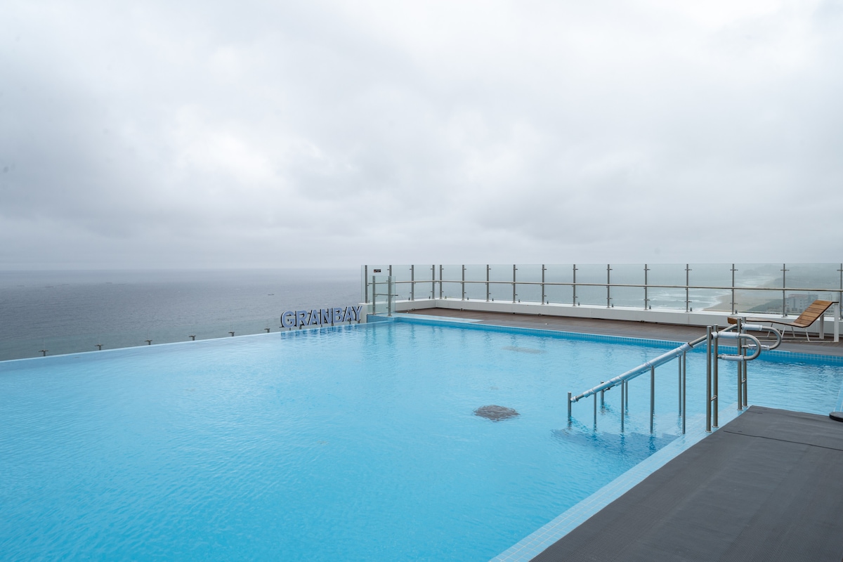 An infinity pool is situated on the 21st floor, featuring a clear blue surface reflecting the cloudy sky. Glass railings surround the pool, offering unobstructed views of the ocean. A sun lounger is placed nearby, providing a spot for relaxation.