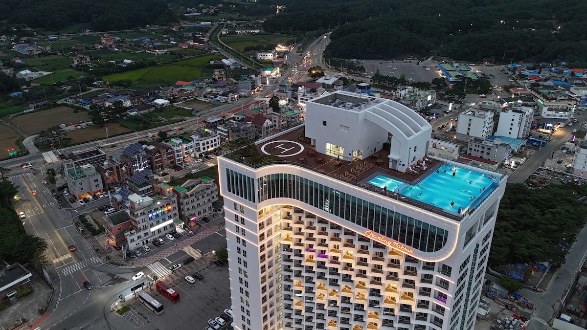 An aerial view captures the Urban Stay building, showcasing its modern architecture and the 21st-floor infinity pool. Surrounding landscapes feature a mix of urban and natural scenery, with roads and green fields visible in the distance.