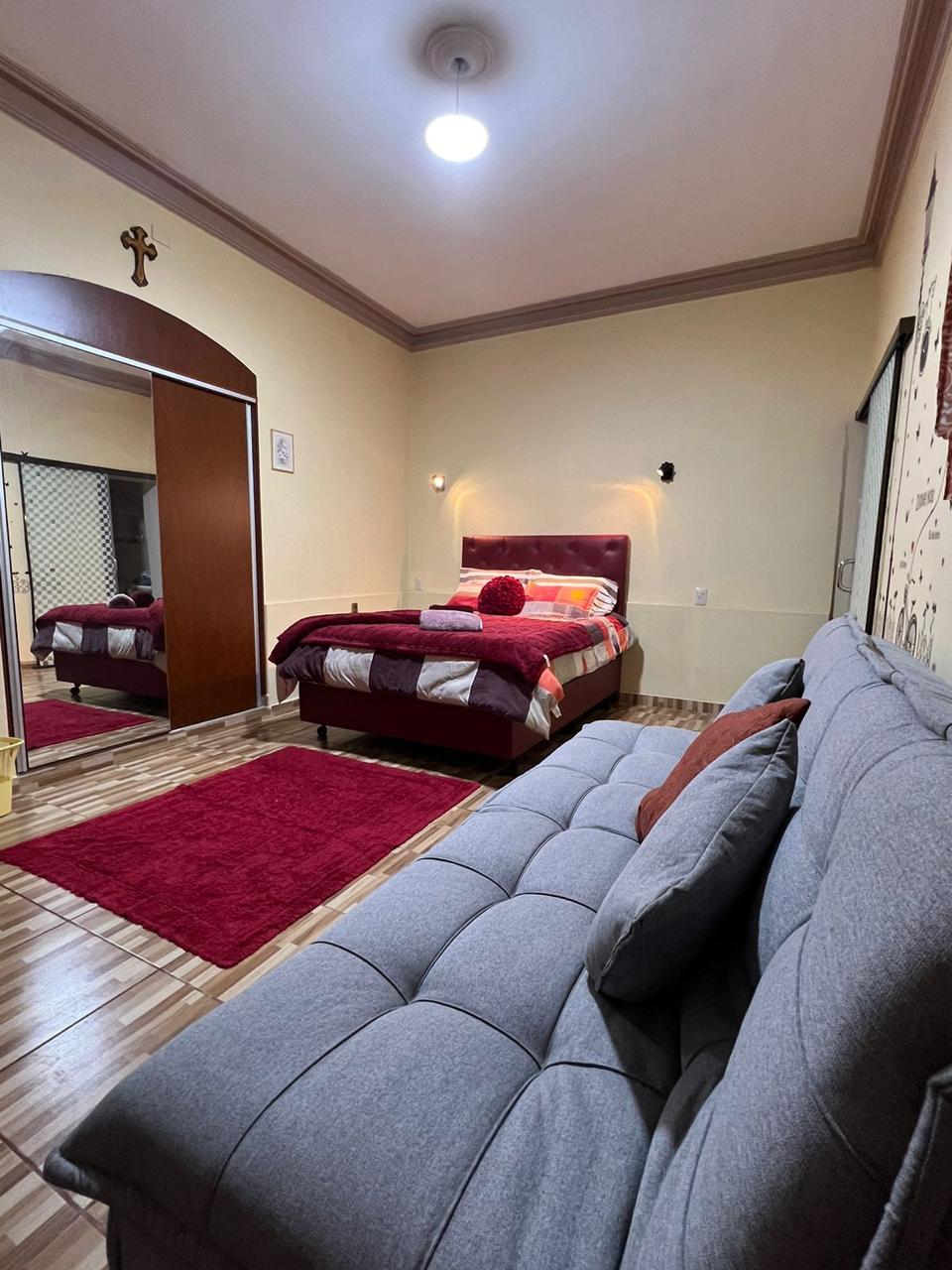 A comfortable double room features a spacious bed topped with decorative pillows. A cozy sofa is positioned nearby, accompanied by a rich red area rug. A large mirror reflects the room's layout, while soft lighting enhances the overall ambiance.