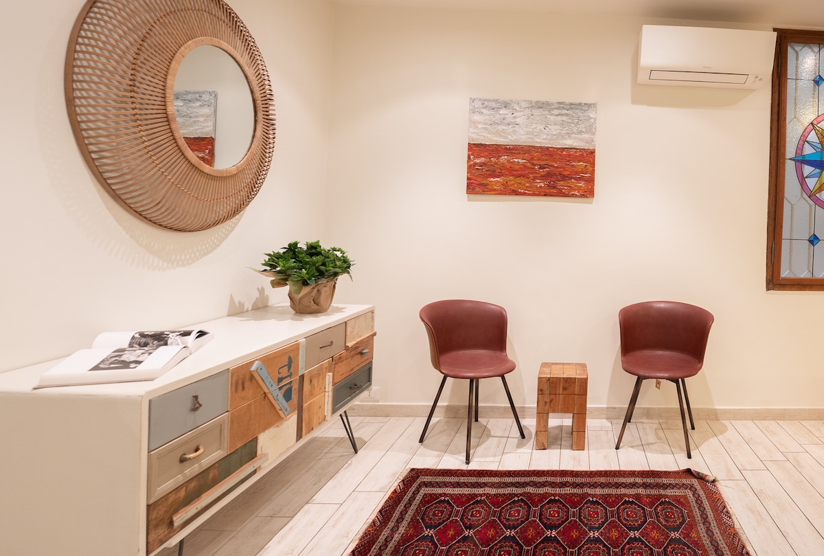 A welcoming entryway is highlighted by a textured circular mirror on the wall, reflecting soft light. Two stylish chairs surround a wooden side table. A vibrant area rug adds warmth to the space, complemented by a small decorative plant and a piece of colorful wall art.