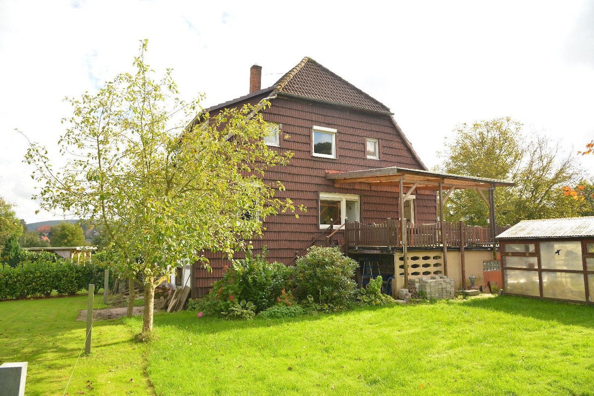 The exterior of a spacious house is shown, featuring brown wooden siding and a covered terrace. A well-maintained garden is visible, along with a tree and shrubbery. Natural light illuminates the scene, creating a welcoming atmosphere.