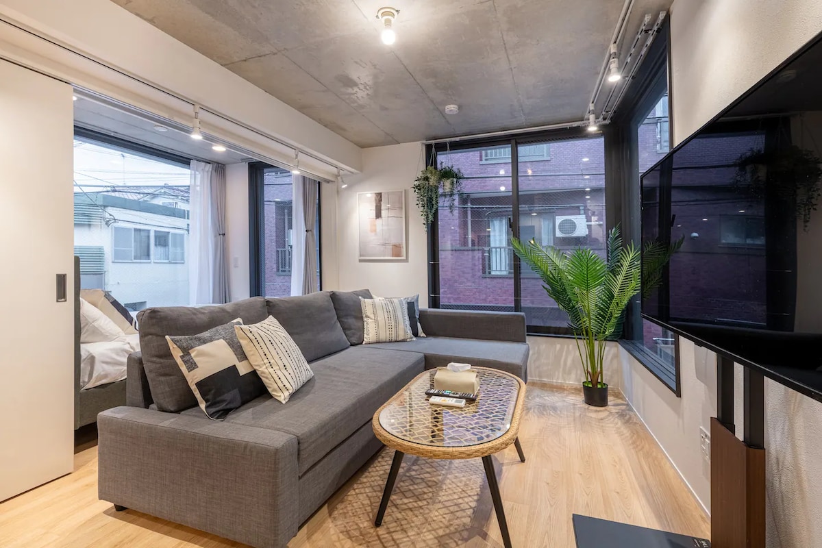 A spacious living area features a gray sofa arranged around a low coffee table. Large windows allow natural light to brighten the space, complemented by indoor plants. A flat-screen TV is mounted on the wall, and textured decor adds visual interest.