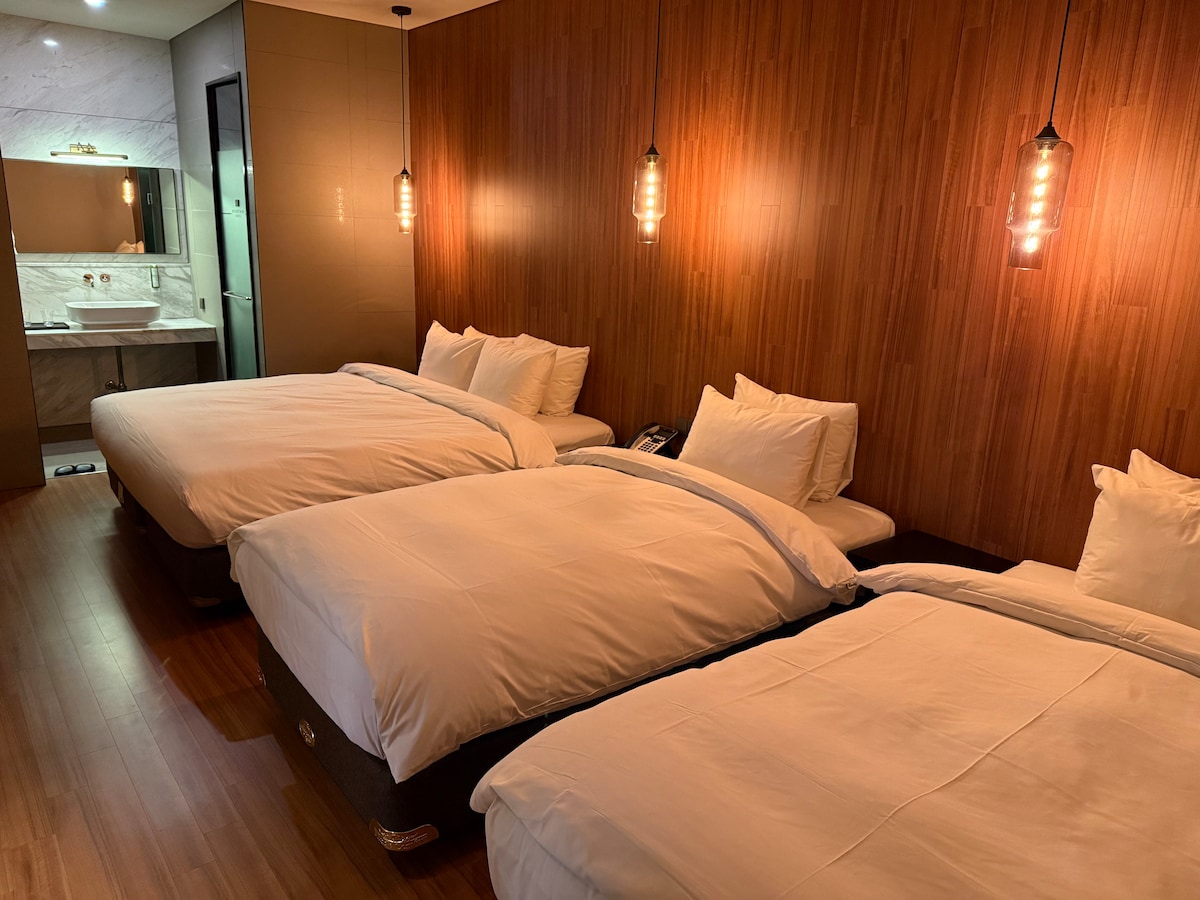 The guest room features three spacious beds with crisp white linens and soft pillows arranged against a wooden accent wall. Soft lighting from pendant fixtures enhances the room's warmth. A sleek bathroom is visible in the background, providing modern amenities.