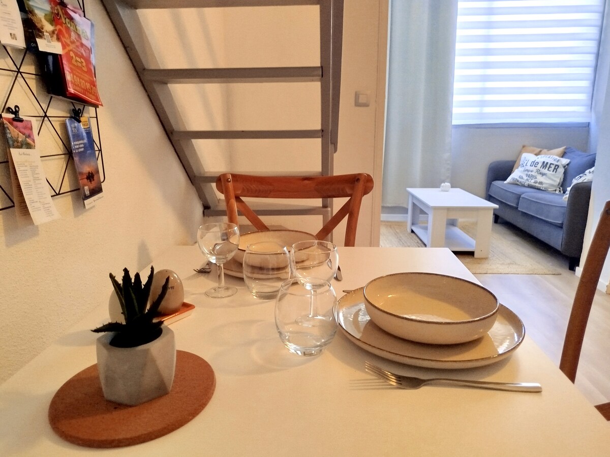 A dining table is set with neutral-colored plates and glassware, accompanied by a small potted plant. In the background, a cozy seating area is visible, furnished with a light-colored couch and a white coffee table. Natural light enters through covered windows.