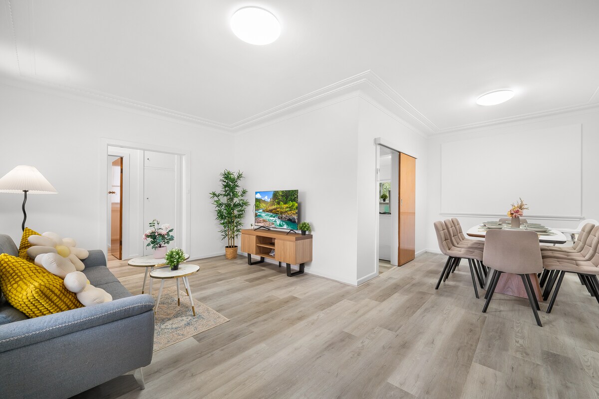 A light-filled living area showcases modern furnishings, including a comfortable sofa and a wooden coffee table with decorative plants. A flat-screen TV is mounted on a TV console, while a dining table with plush chairs is positioned nearby, allowing for a cohesive social space.