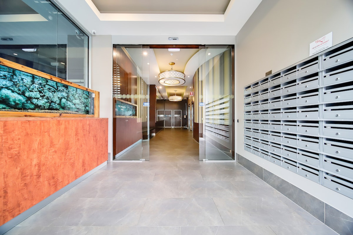An entryway is marked by glass double doors, leading into a modern lobby. To the side, rows of secure mailboxes are visible, with a decorative wall featuring textured panels. Soft lighting highlights the spacious area, providing an inviting first impression.