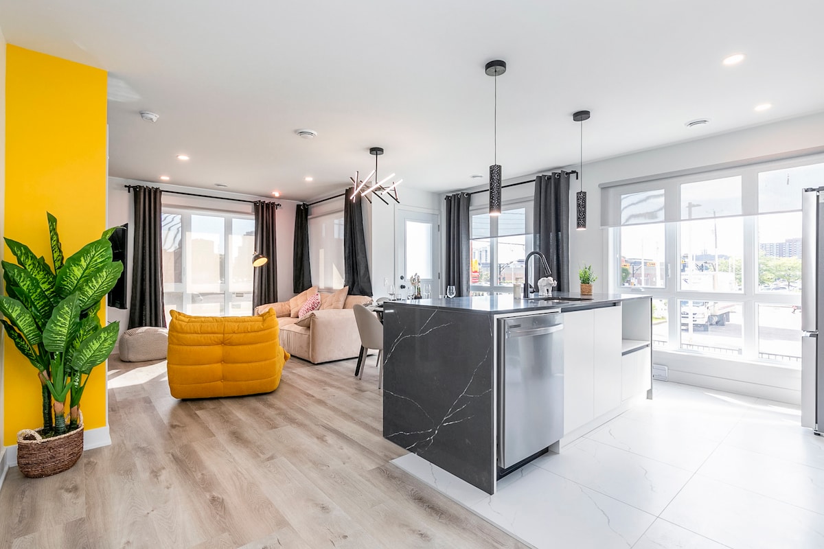 An open-concept living space features large windows allowing natural light, with a bright yellow accent wall. A modern kitchen island with bar seating is visible, accompanied by a comfortable seating area and a decorative indoor plant. Soft furnishings add a welcoming touch.
