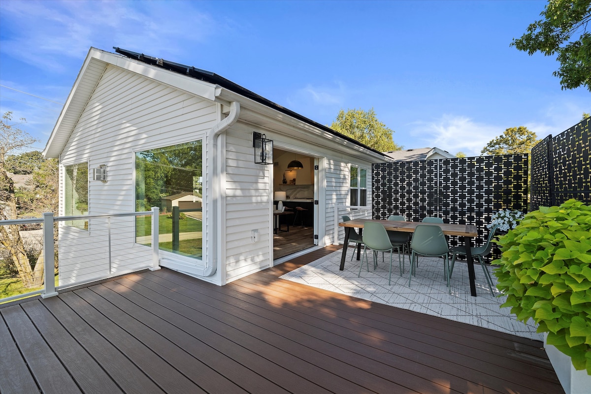 An outdoor dining area is situated on a spacious deck, featuring a wooden table surrounded by modern chairs. Large glass doors lead to the interior, enhancing the connection between indoor and outdoor spaces. Lush greenery and patterned privacy screens add to the serene atmosphere.