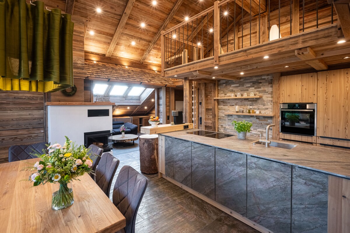 A modern kitchen area with wooden cabinetry and stone accents is highlighted. An island with a built-in sink and seating is visible, along with a dining table adorned with a flower arrangement. Warm lighting enhances the rustic wooden beams and walls, creating a cozy atmosphere.