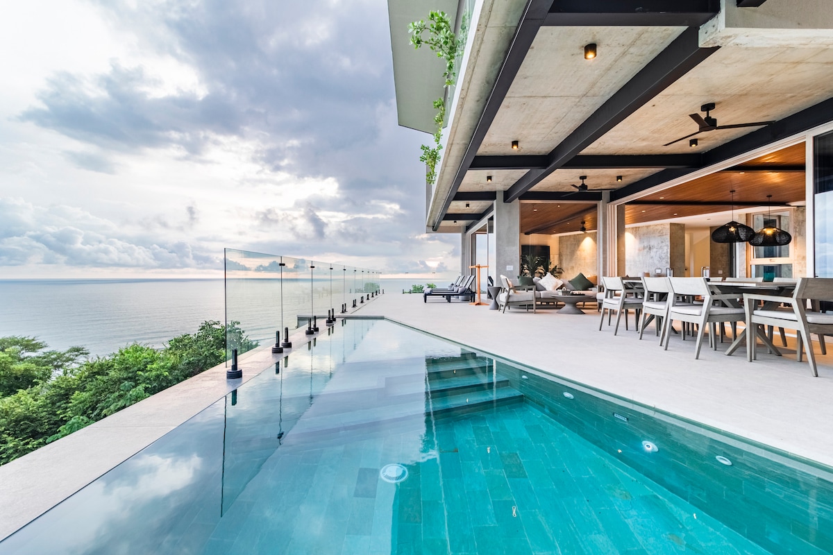 A modern outdoor space features an infinity pool reflecting the sky, surrounded by glass railings. The terrace includes a large dining table and comfortable seating arrangements, offering expansive views of the ocean and surrounding landscape under a soft, cloudy sky.