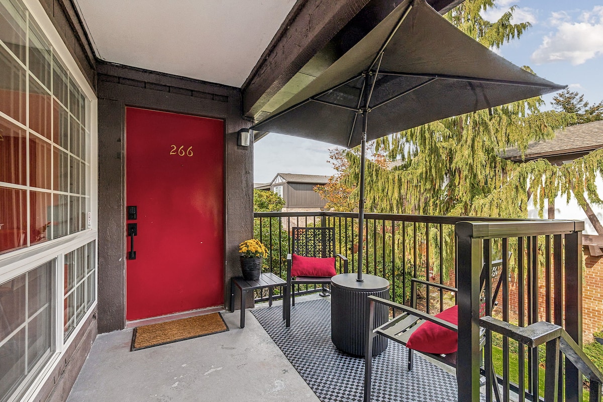 An inviting outdoor balcony features two chairs with red cushions and a round table, designed for relaxation. A large umbrella provides shade, while a small potted plant adds a touch of greenery. The bright red door serves as a vibrant focal point against the neutral exterior.