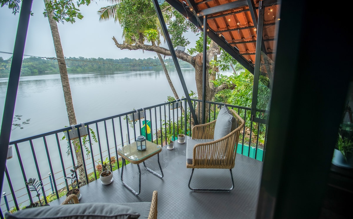 A serene balcony overlooking a calm lake is furnished with rattan chairs and a coffee table. Potted plants add a natural touch, while fairy lamps provide a warm glow. The lush greenery surrounding the area enhances the tranquil setting, perfect for relaxation.