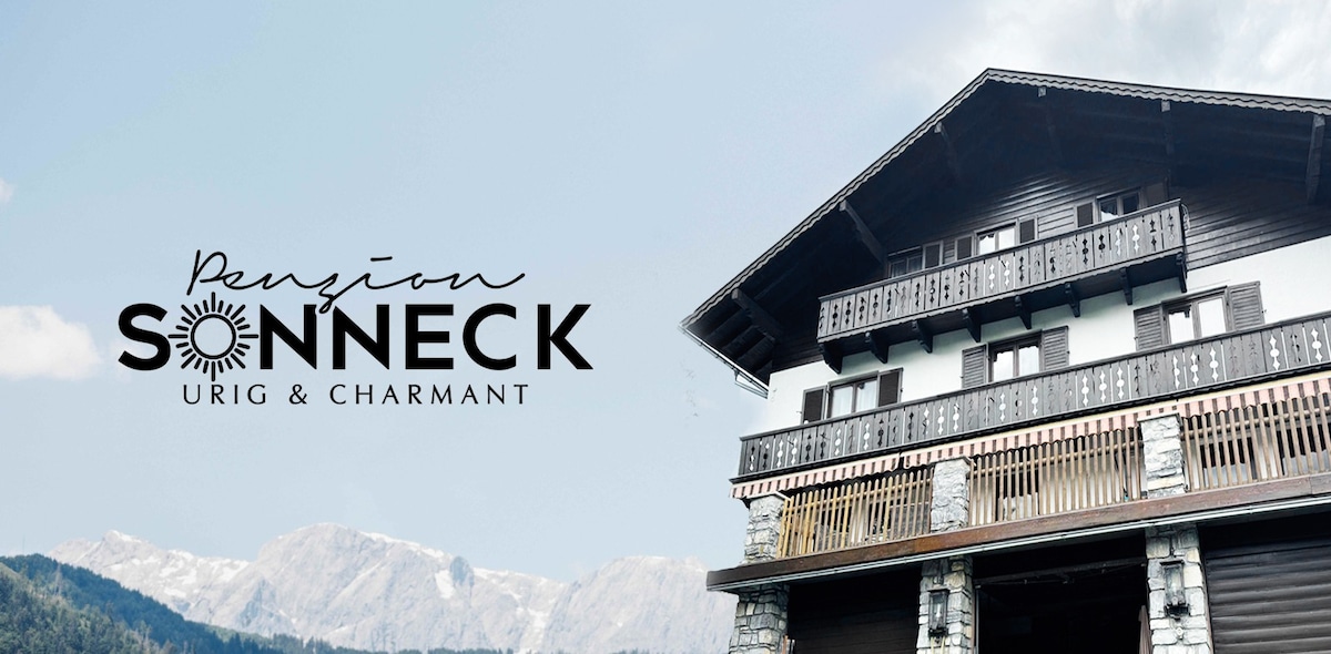 A charming alpine building features a traditional wooden facade with multiple balconies. Sunlight reflects off the stone accents, creating a warm and inviting appearance. Majestic mountains form a backdrop, enhancing the picturesque setting.