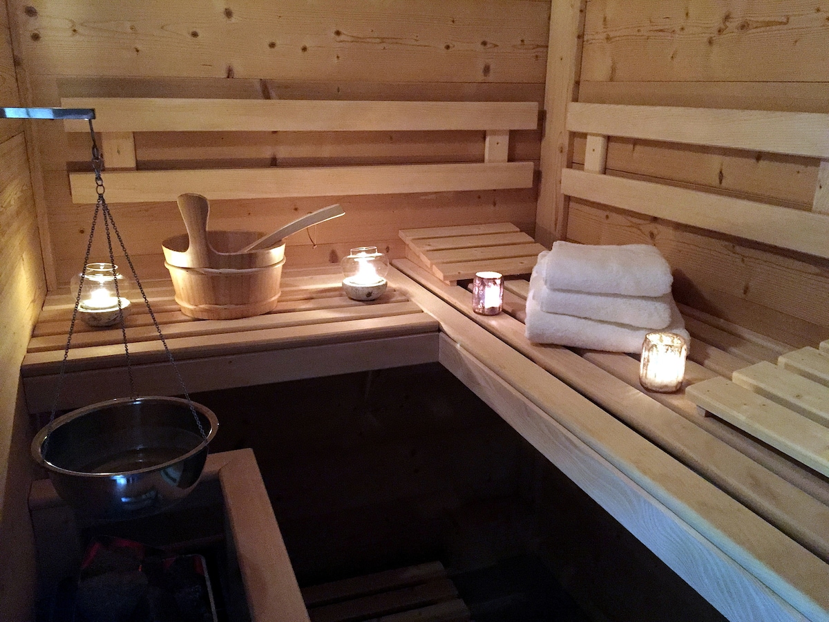 A warm sauna interior is crafted from light wood, featuring benches on either side. Softly glowing candles and neatly stacked towels create a serene atmosphere. Traditional sauna accessories, including a wooden bucket and ladle, are placed beside a metal container for water.