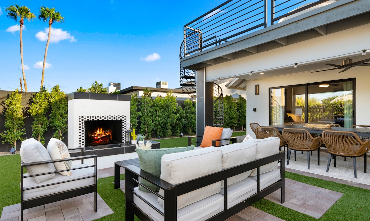An outdoor living area is highlighted by a spacious seating arrangement, featuring a sectional sofa and cozy chairs. A built-in fireplace adds warmth, and a dining table can be seen in the background. Lush greenery and clear blue skies create a serene atmosphere.