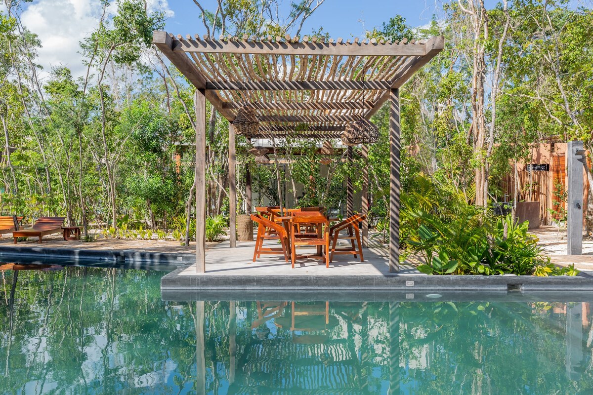 An outdoor dining area is positioned beside a serene pool, featuring a wooden table surrounded by matching chairs. A pergola above offers partial shade. Lush greenery and trees create a natural backdrop, enhancing the peaceful ambiance of the space.