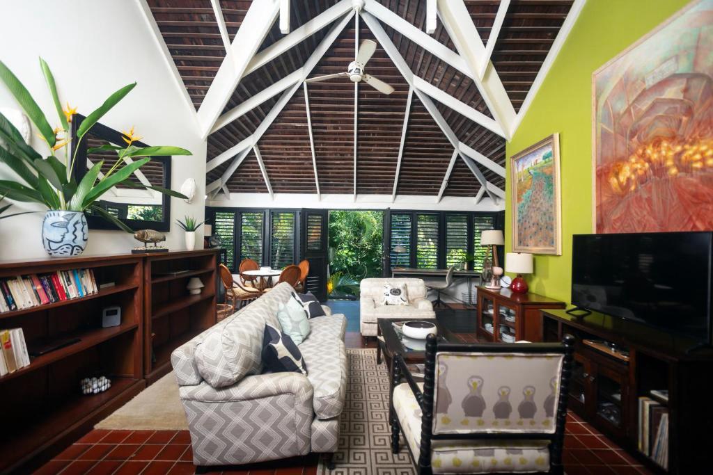 The central space features a vaulted ceiling with open rafters, surrounded by jalousie windows that offer garden views. A comfortable seating area includes a patterned sofa and armchair, with a television and bookshelf nearby. Bright artwork adorns the green accent wall, enhancing the lively ambiance.