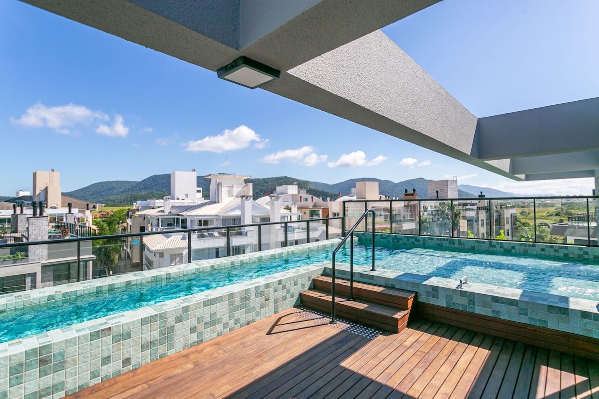 A spacious rooftop area features a glistening pool surrounded by tiled decking. The scene includes a panoramic view of the skyline and distant mountains under a clear blue sky, inviting relaxation and enjoyment.