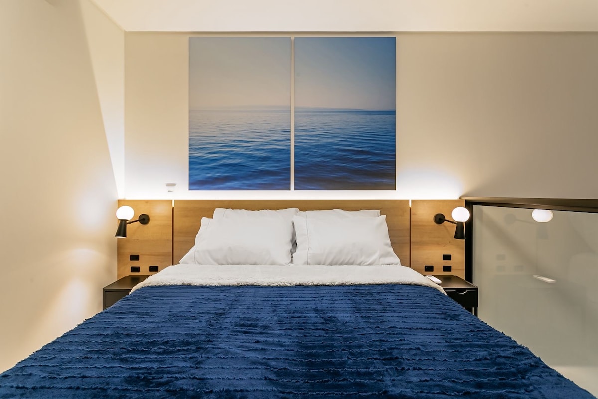 A cozy sleeping area features a spacious bed dressed in a soft blue blanket. Two serene artworks of the ocean hang above the headboard, while contemporary bedside lamps provide soft lighting. The warm tones of the wood accent the inviting atmosphere.