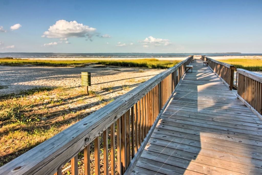 A wooden boardwalk stretches into the distance, leading to the sandy beach. Flanking the path, grass and natural vegetation are visible. The serene sky features scattered clouds, and a water view can be seen beyond the beach area, adding to the tranquil landscape.