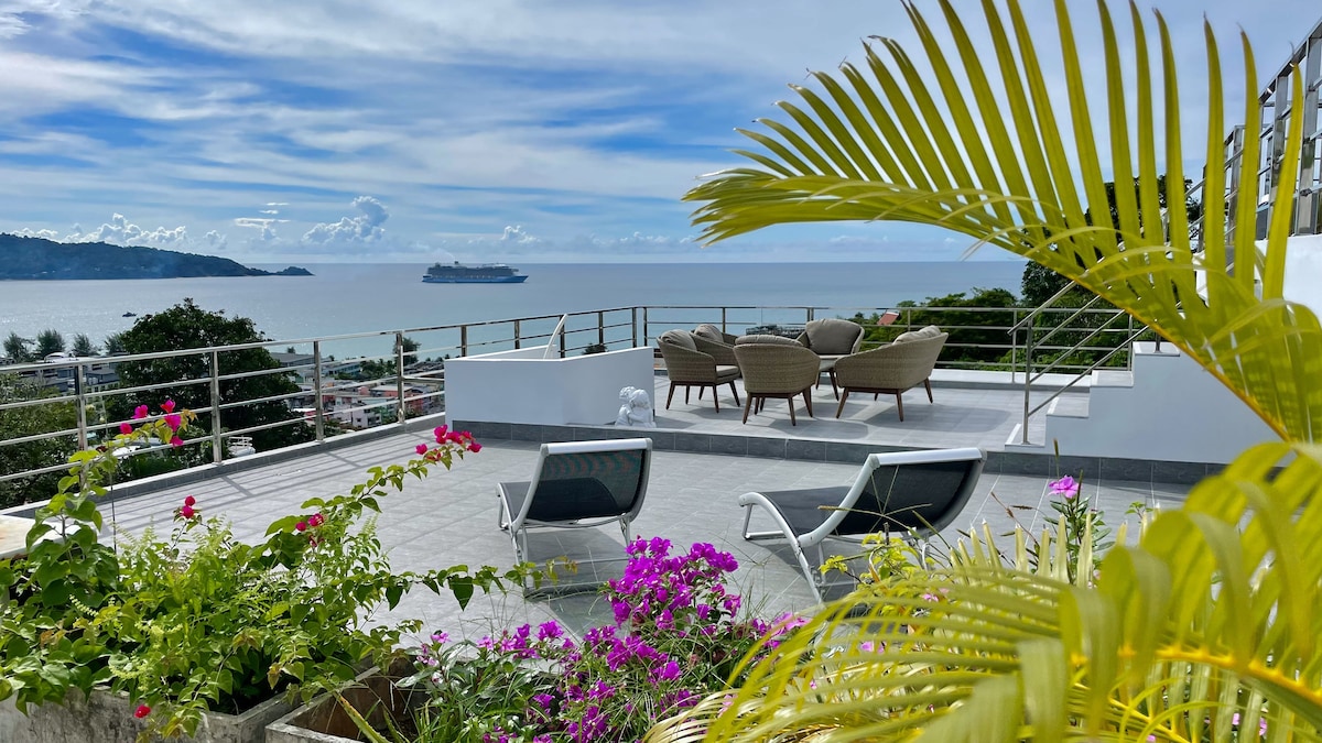 A spacious rooftop terrace features comfortable seating arrangements overlooking the sea. Lush potted plants with vibrant flowers add color to the area, while the panoramic views of the ocean and distant landscape provide a serene backdrop.
