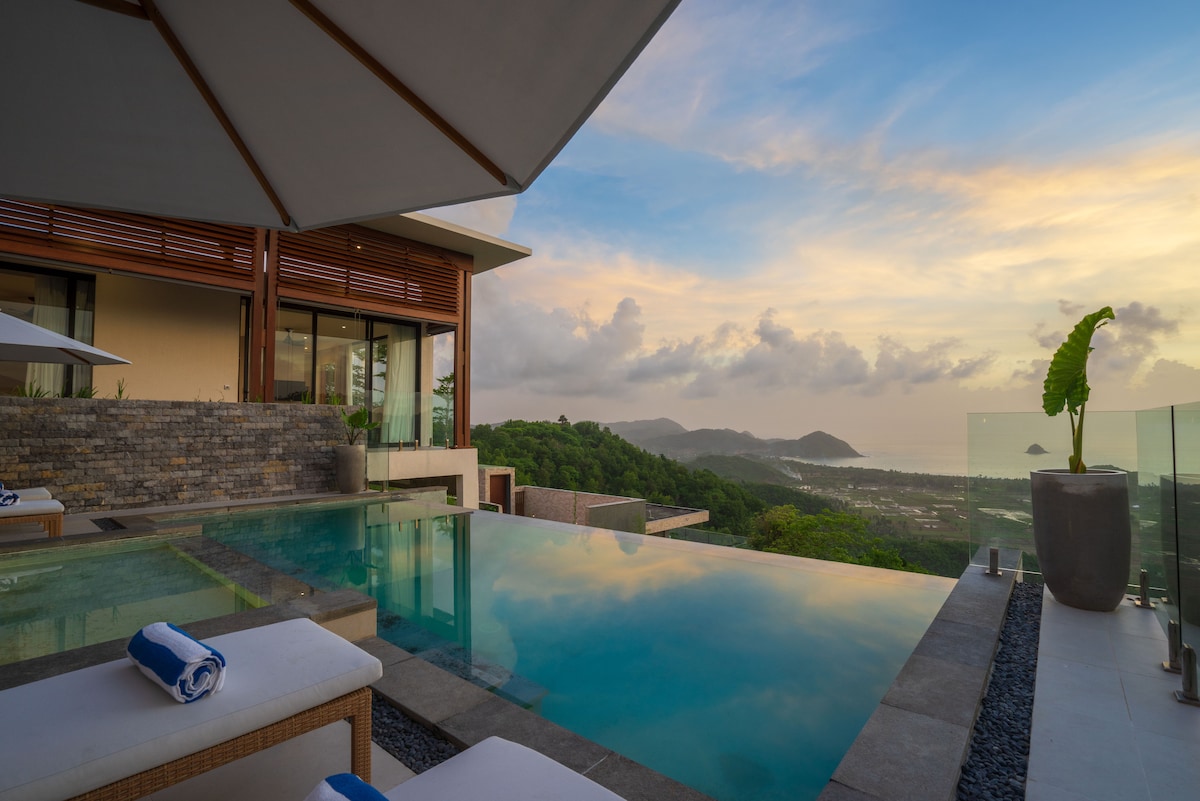 The private infinity pool extends towards the horizon, showcasing stunning ocean views. Sun loungers are positioned beside the pool, accompanied by large umbrellas for shade. Large glass doors lead to the villa, with lush green hills visible in the background against a colorful dusk sky.