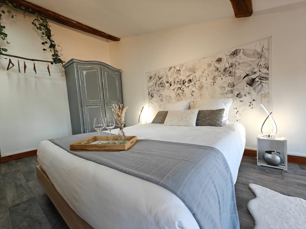 A spacious bedroom features a comfortable double bed with decorative pillows and a light-colored blanket. A wall art piece with abstract designs serves as a backdrop. A wood-paneled ceiling adds character, while soft lighting is provided by two bedside lamps. A decorative tray with glasses rests on a side table.
