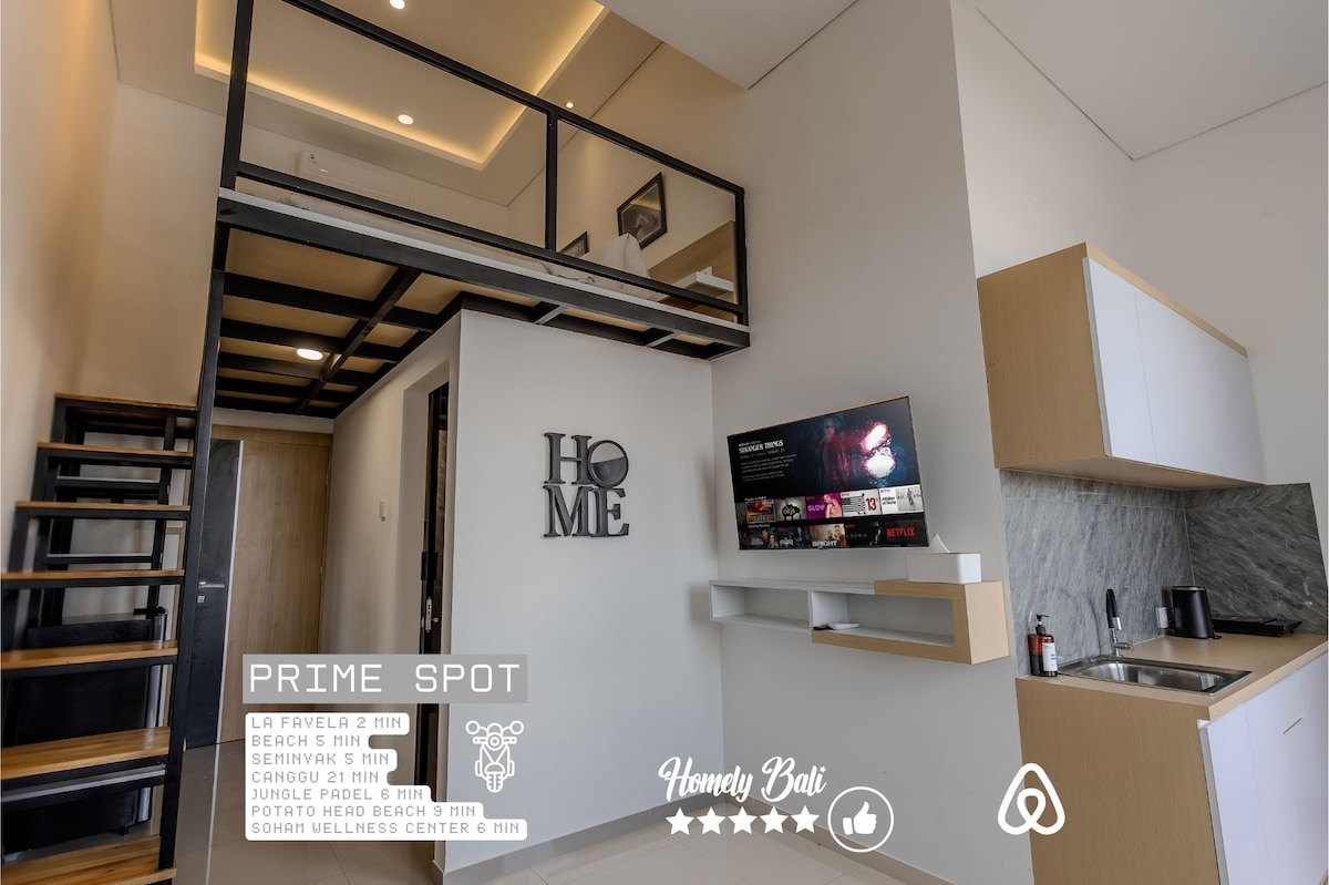 An airy studio space features a modern mezzanine level, showcasing an open-plan design. A sleek kitchen area is visible, accompanied by a wall-mounted television. A spiral staircase leads to the upper loft area, while a bright, neutral color palette enhances the contemporary feel.