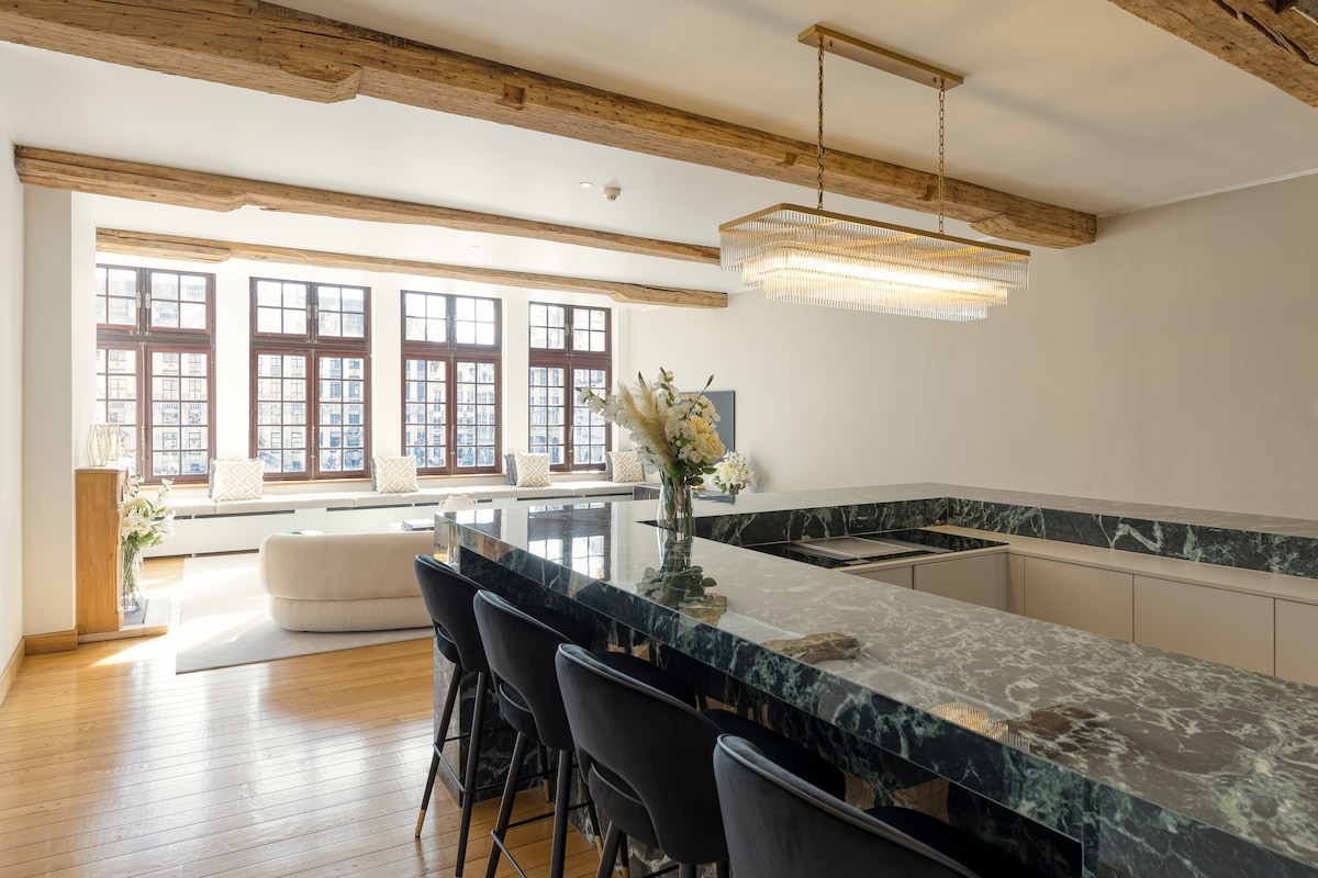 A modern kitchen features a dark marble countertop with seating for six at black, upholstered chairs. Above, a chandelier with crystal accents illuminates the space. Large windows allow natural light to fill the room, highlighting the wooden beams across the ceiling.