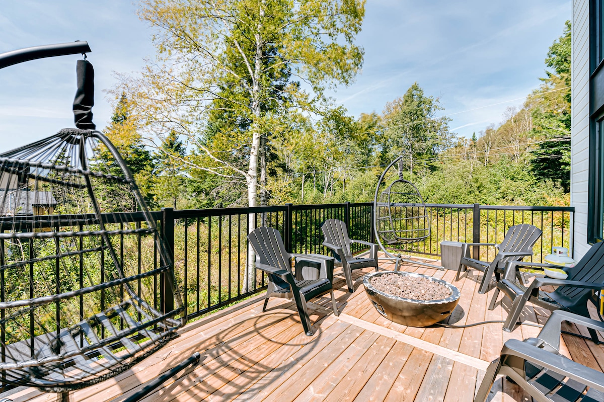 An outdoor terrace features a circular fire pit surrounded by multiple Adirondack chairs. Two hanging chairs are positioned on the side, along with a backdrop of lush green trees, offering a serene natural setting.