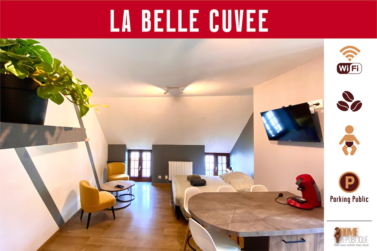 The interior space features a cozy layout with a spacious double bed and a small table with two chairs. A coffee maker sits on a surface near the wall. Light enters through two windows, illuminating the neutral-toned walls and wooden flooring.