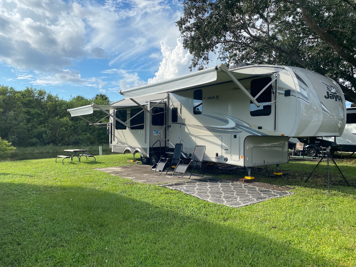 A spacious fifth wheel camper is parked on a grassy lot, featuring two retractable awnings. A patio area is furnished with four lounge chairs, and a picnic table is positioned nearby. Lush greenery surrounds the site, with a blue sky and scattered clouds overhead.