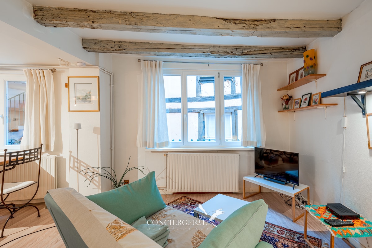 The living area features exposed wooden beams and large windows allowing natural light to fill the space. A comfortable sofa and a small television provide a cozy seating area. Shelves display decorative items, enhancing the inviting ambiance.