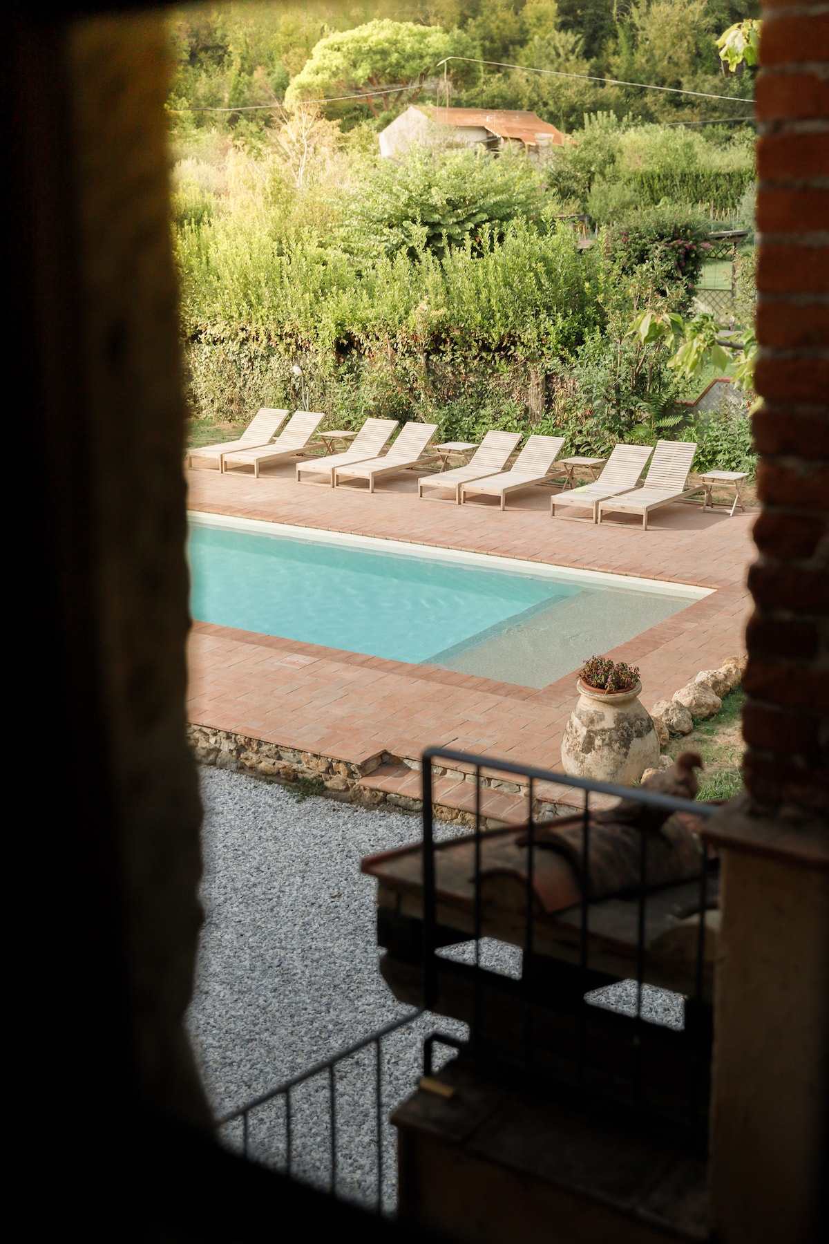 A view of the salt-water pool is framed by a stone archway. Several lounge chairs are arranged neatly on the patio beside the pool, surrounded by lush greenery. The tranquil atmosphere is enhanced by the natural setting.