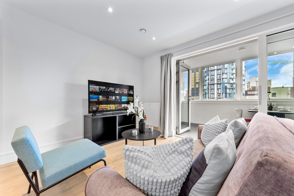 A living area is presented with a plush sofa and a blue accent chair. A flat-screen TV is positioned on a sleek black unit, while a small coffee table is adorned with decorative items. Large windows allow natural light to fill the space, with views of the city outside.